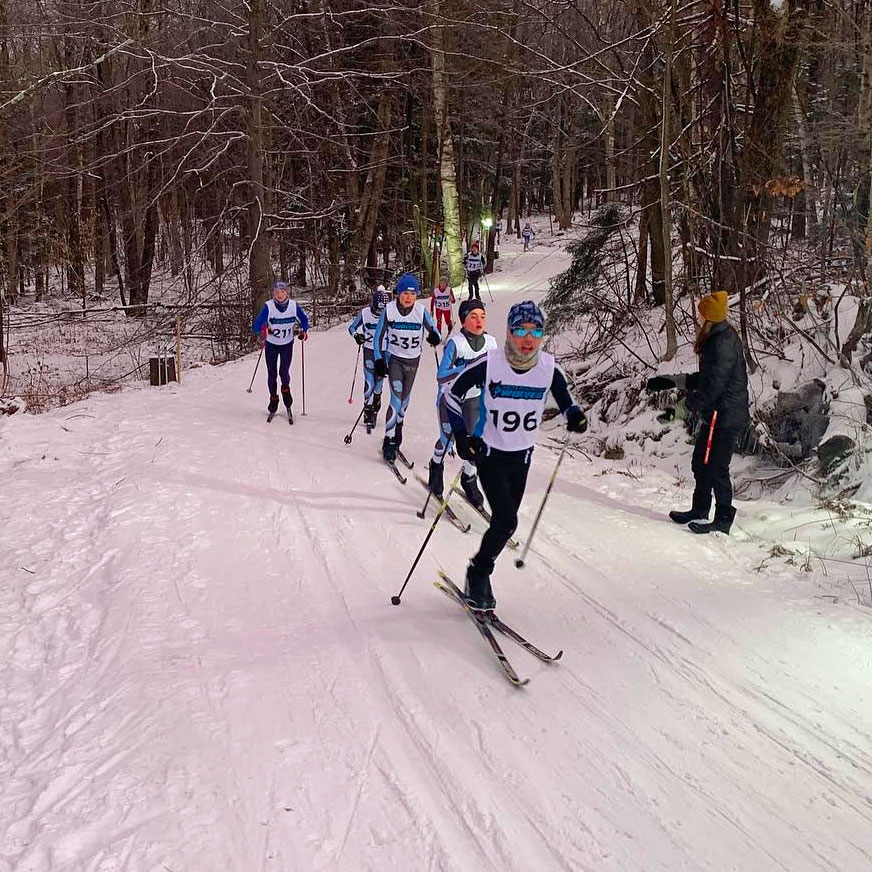 Cross-country skiers racing on a snow-covered track in a wooded area in Vermont.