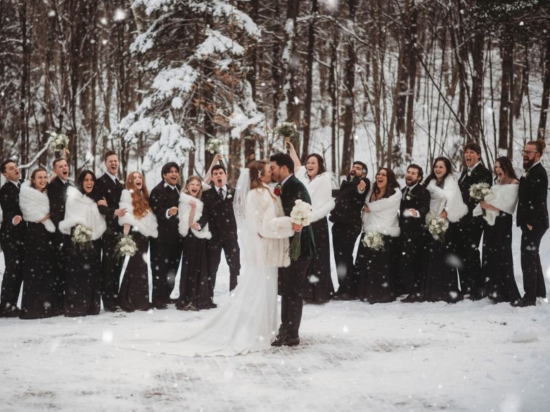 A wedding couple shares a kiss amidst a snowy Vermont scene, surrounded by a cheerful bridal party.
