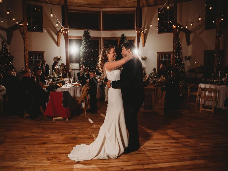 A couple sharing their first dance at a Vermont barn wedding reception with guests seated around them and festive decorations in the background.